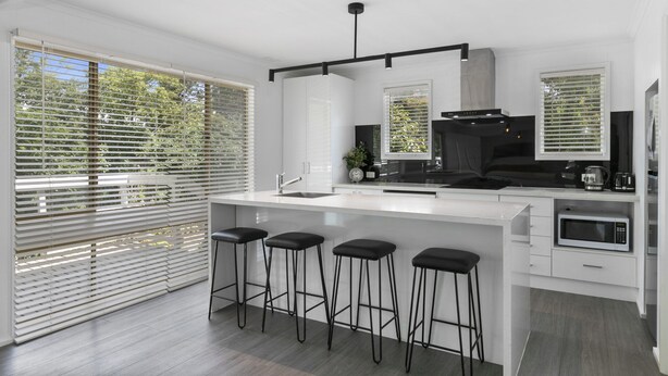 A modern kitchen with a large island, bar stools, a built-in oven, and a window with blinds. - Beauty AND THE Peace - Inlet Side (Inverloch)