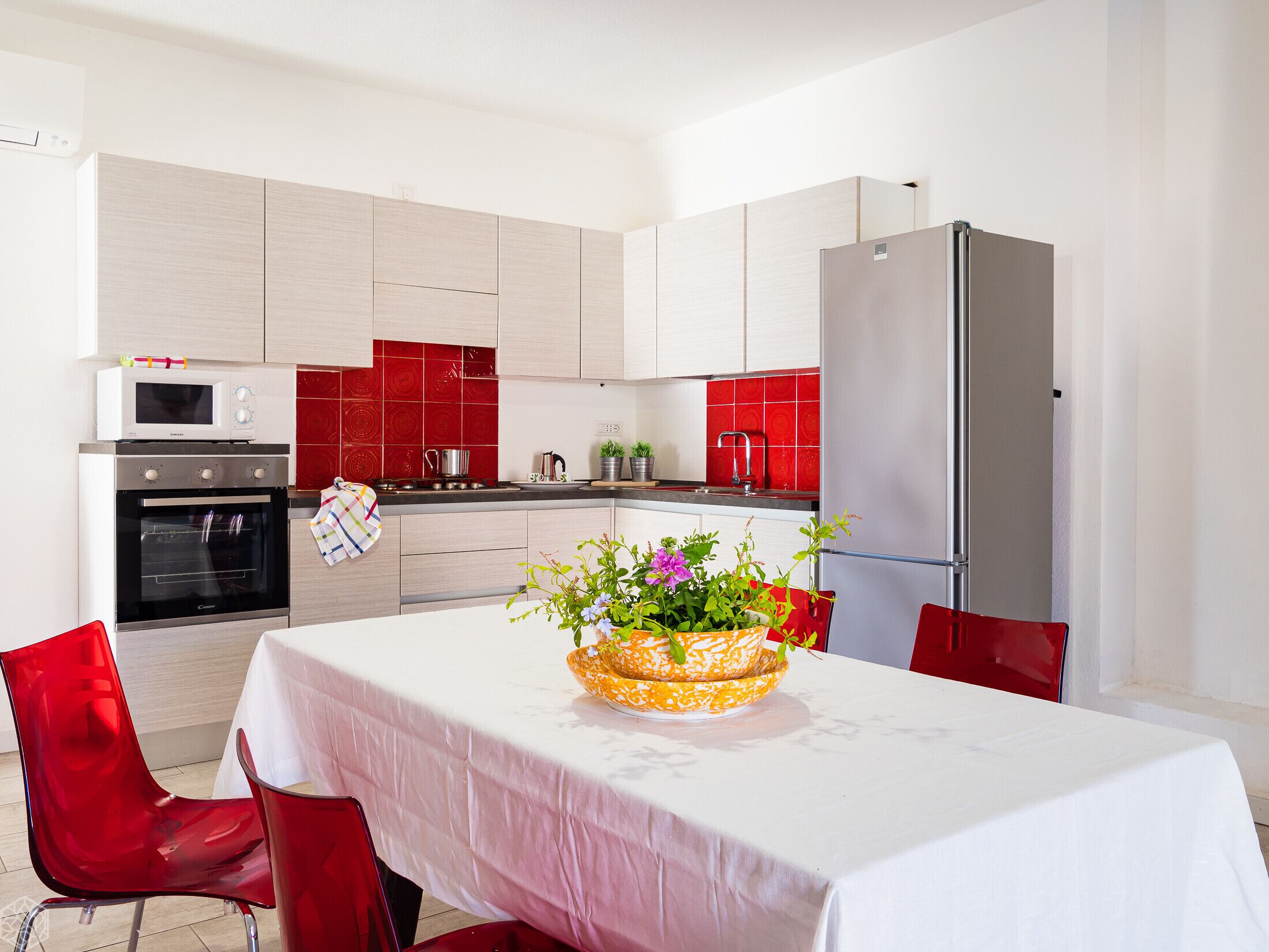 A modern kitchen with white cabinets, a red backsplash, a microwave, an oven, a sink, and a refrigerator. There is a table with a white tablecloth and a basket of flowers.