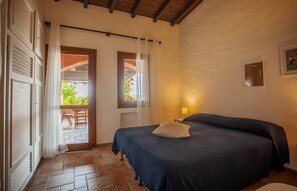 Ein Schlafzimmer mit Bett, Nachttisch, Lampe, Fenster mit Vorhängen und Blick ins Freie.