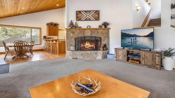 A spacious living room with a stone fireplace, wooden furniture, a television, and a large window overlooking a lake. - 13 Modoc-Sunriver, Oregon: Hot Tub, BBQ, WiFi, Ping Pong, Foosall, SHARC Aquatics, Tubing Hill, Mt. Bachelor, Golf, Deschutes River, Tennis, Bike Paths (Sunriver)