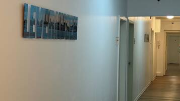 A hallway with white walls, a wooden floor, and a ceiling light. There is a framed artwork on the wall.
