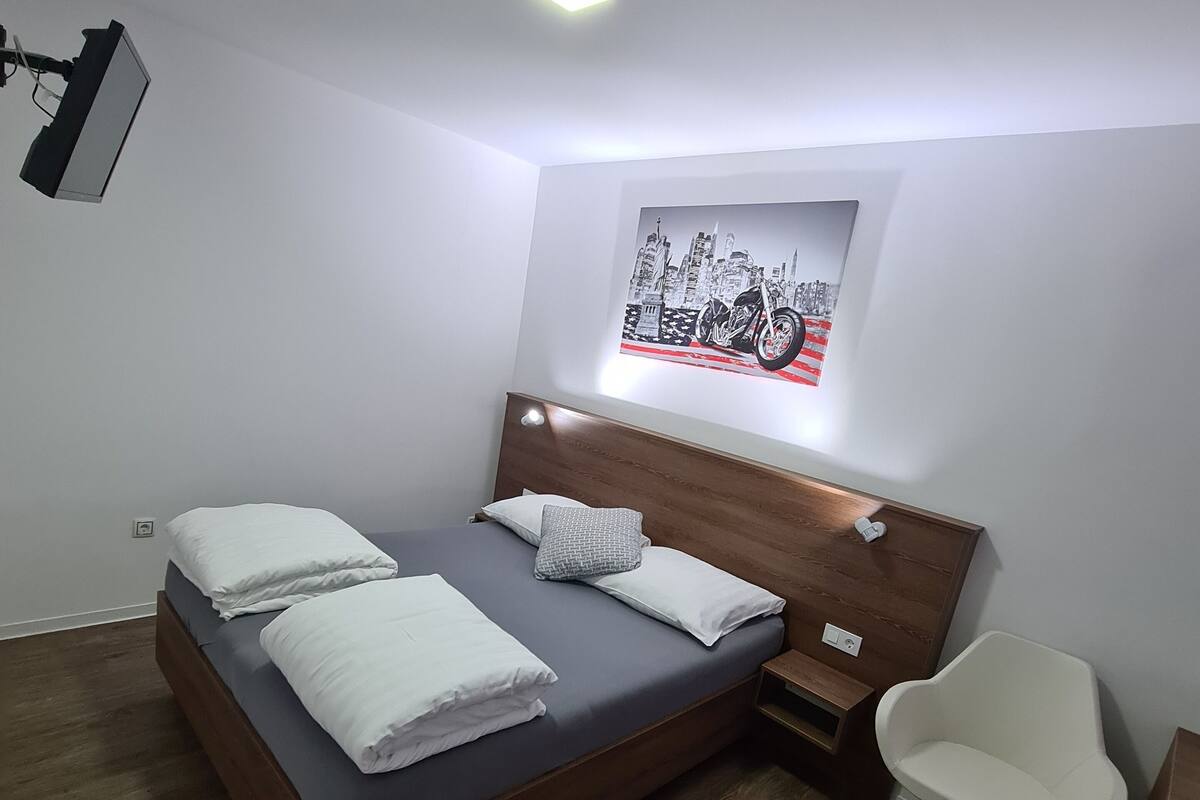 A modern bedroom with a wooden bed, white pillows, a headboard with built-in lighting, a wall-mounted cityscape artwork, and a white chair.