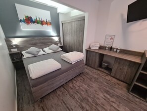 Une chambre d’hôtel moderne avec un grand lit, une tête de lit en bois, une table de chevet avec une lampe, un miroir, un bureau en bois avec une serviette et des articles de toilette, et un panneau encadré accroché au mur.