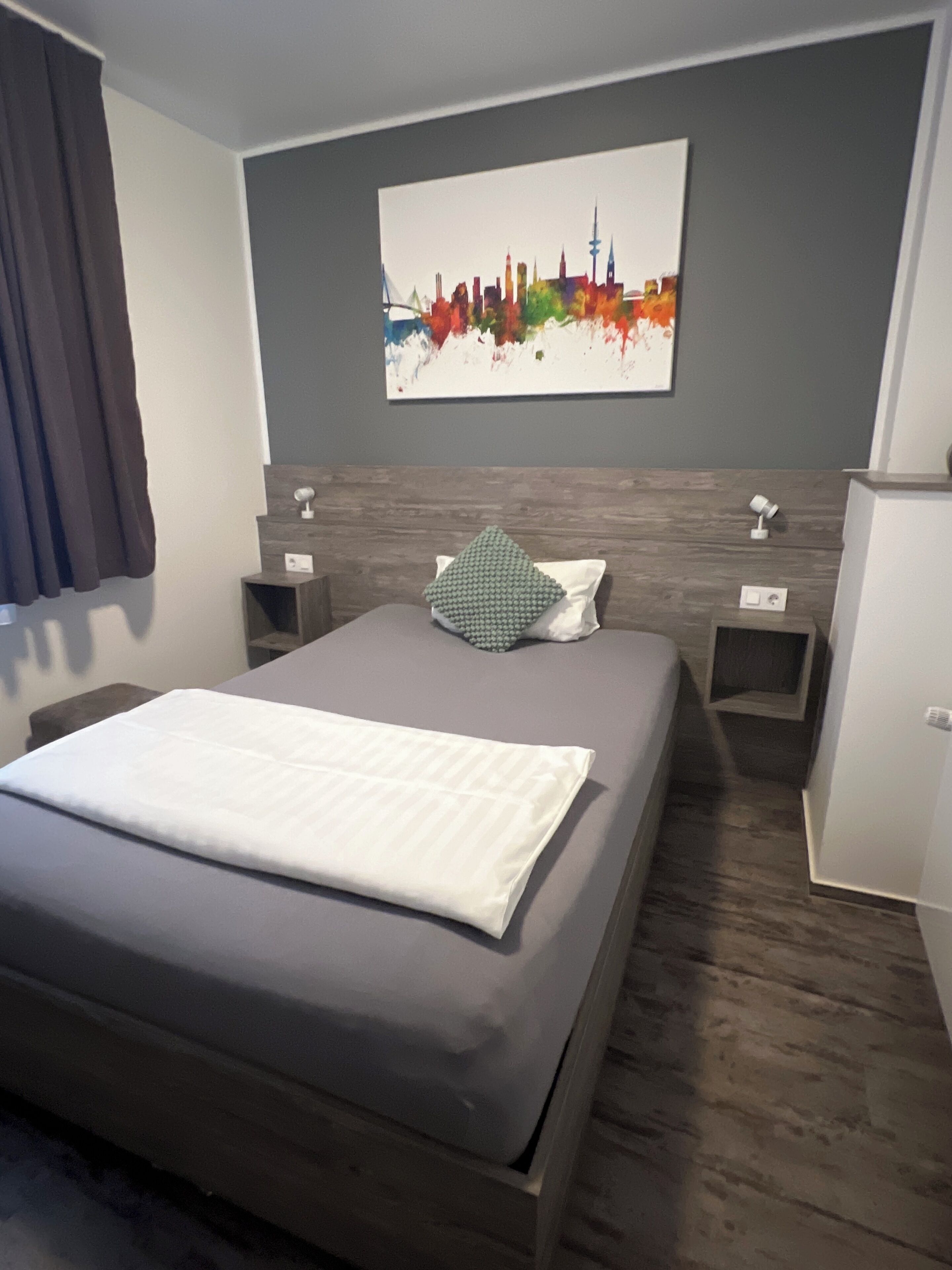 A modern hotel room with a large bed, a cityscape painting on the wall, and a small bedside table with a lamp.