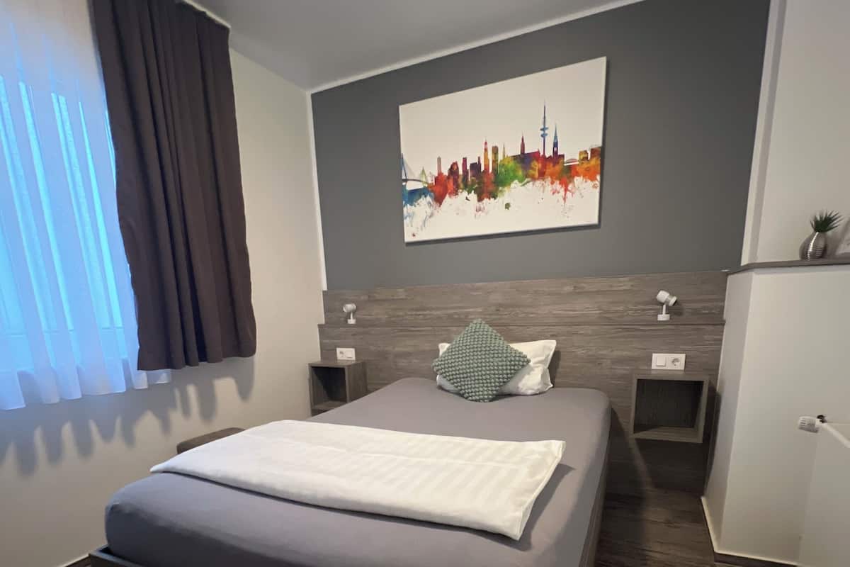A modern bedroom with a bed, a nightstand, a dresser, a window with curtains, and a city skyline painting on the wall.