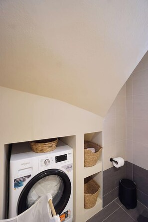 Uma lavanderia compacta com máquina de lavar Whirlpool de abertura frontal, cestos para armazenamento e uma lixeira.