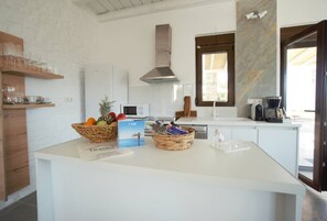 A modern kitchen with white cabinetry, a central island with baskets of fruit, a microwave, and a stove. - Sea Horizon Villa Ikaros by DomisiGroup (Ag. Triada)