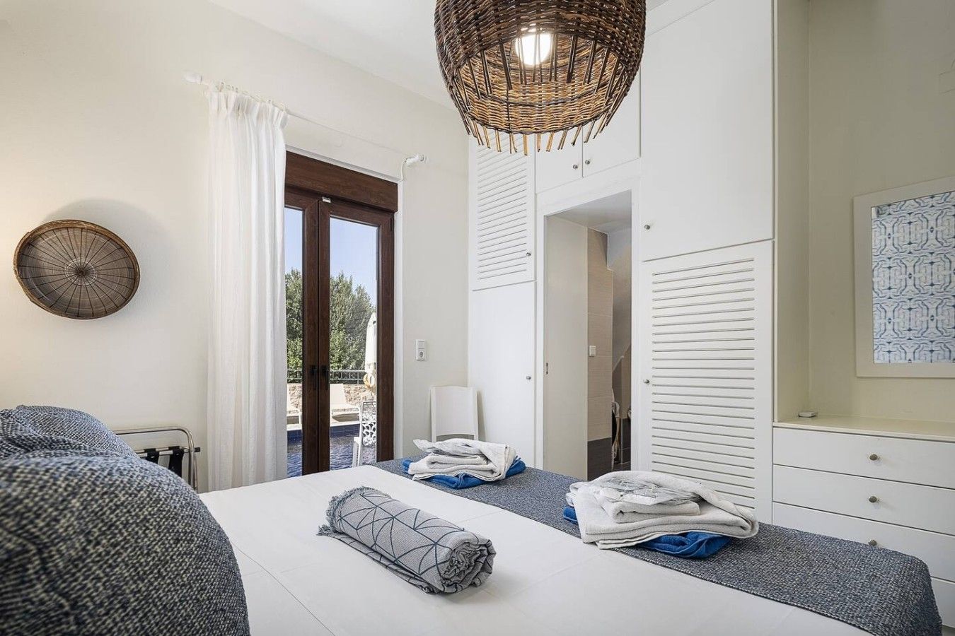A hotel room with a bed, a bedside table, a hanging light fixture, a view through a door, and a wall-mounted fan.