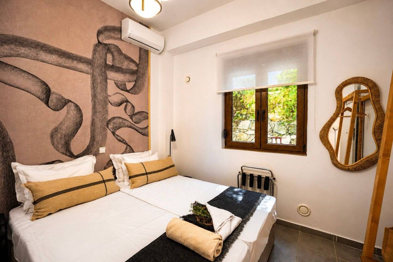 A bedroom with a large bed, a window with a view, a mirror, and an air conditioning unit.