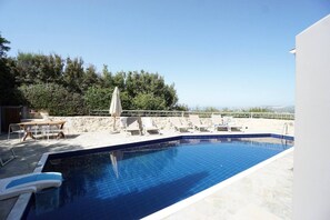 A swimming pool with lounge chairs and a table, surrounded by a white wall and a view of the sea. - Sea Horizon Villa Ikaros by DomisiGroup (Ag. Triada)