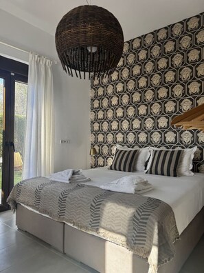 A bedroom with a bed, a hanging light fixture, patterned wallpaper, and a view through a window. - Sea Horizon Villa Ikaros by DomisiGroup (Ag. Triada)
