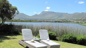 Deux fauteuils de jardin en osier avec des coussins, une bouteille et un verre posés sur une petite table, donnant sur un lac avec des montagnes en arrière-plan.