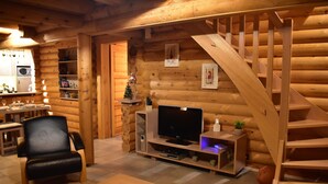 A log cabin interior with a wooden staircase, a television on a wooden stand, a dining table with chairs, and a kitchen area. - La Bresse Lodge - Air-Conditioned Log Cabin (La Bresse)