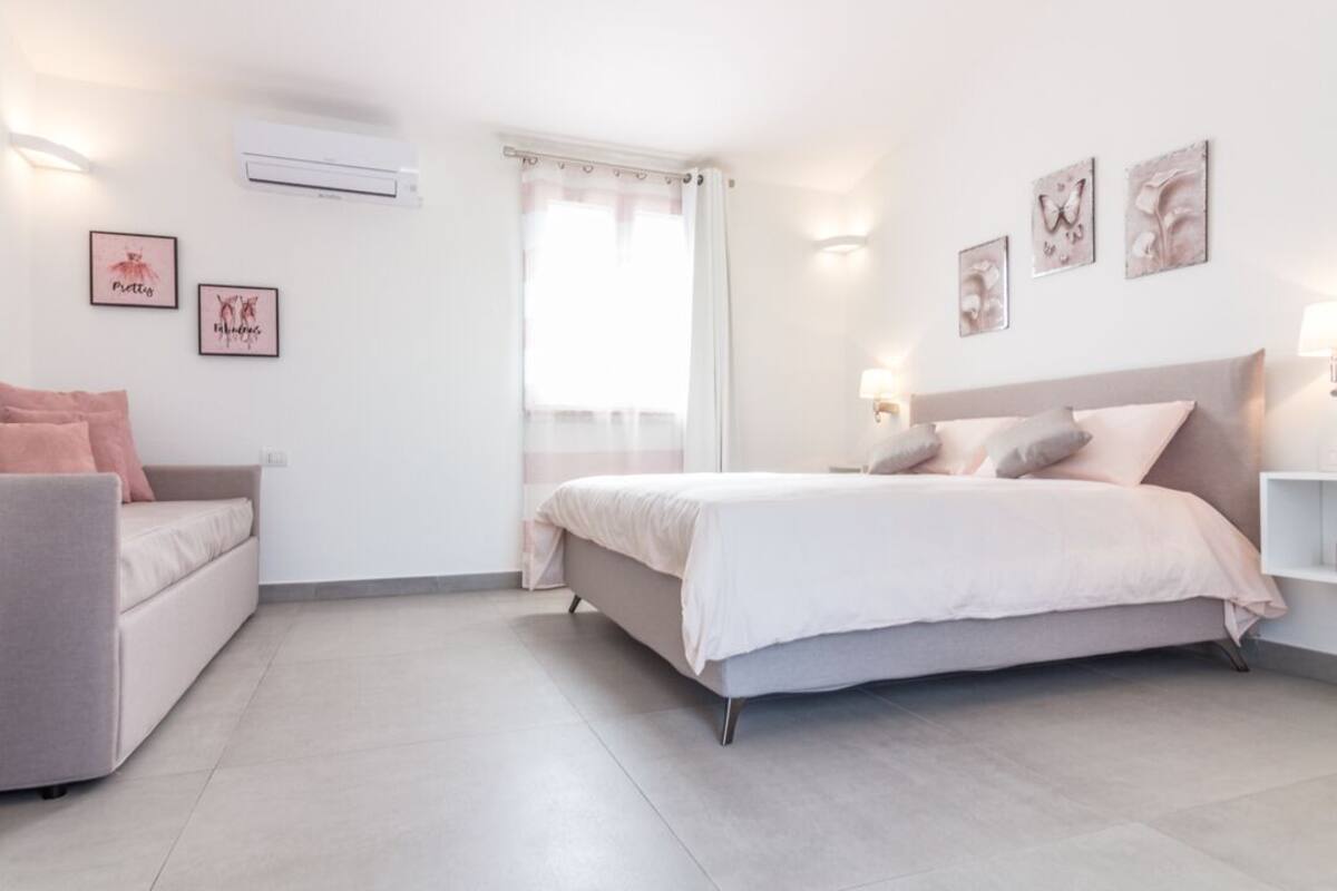 Een slaapkamer met een bed, een bank en een raam met gordijnen. Er zitten wandlampen en ingelijste foto's aan de muren.