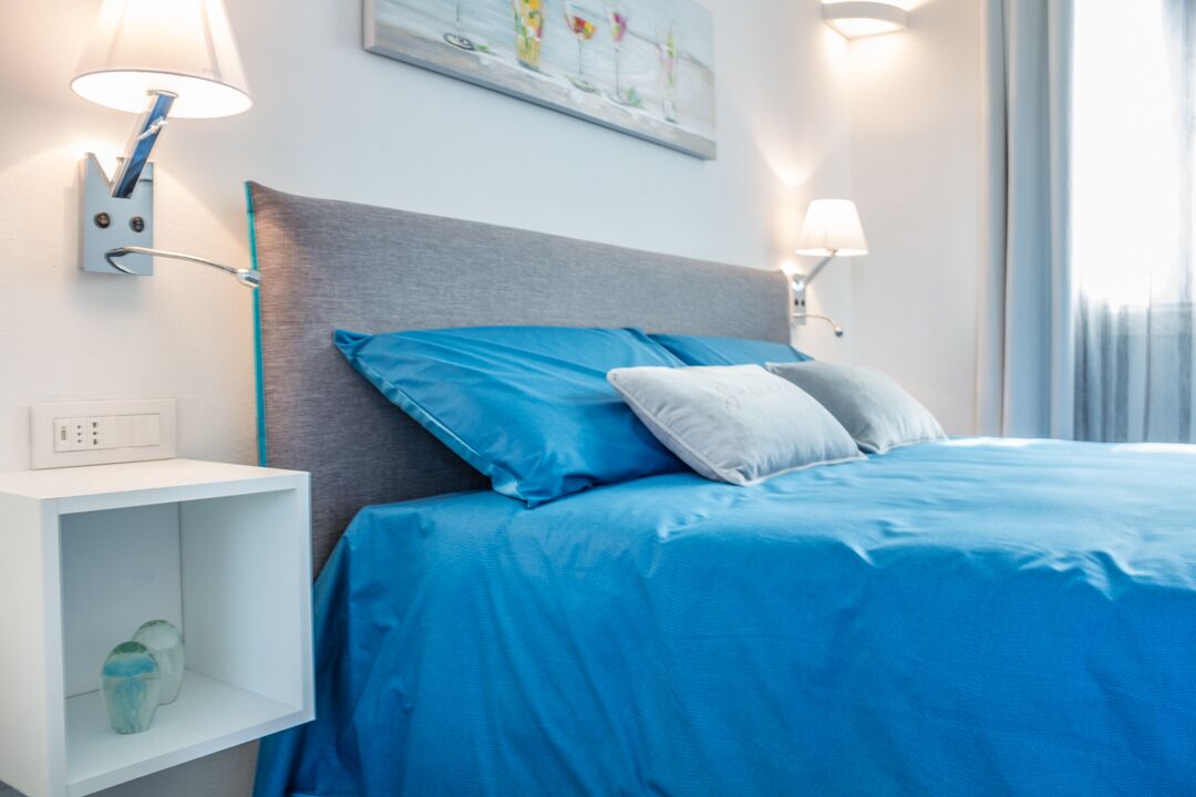 A modern hotel room with a bed, two wall-mounted lamps, a shelf with decorative items, and a window with curtains.