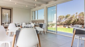 Een moderne eethoek met witte stoelen en tafels, grote ramen en uitzicht op de omgeving.