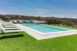 Piscina rectangular con borde blanco, rodeada por un césped cuidado y sillas reclinables blancas.