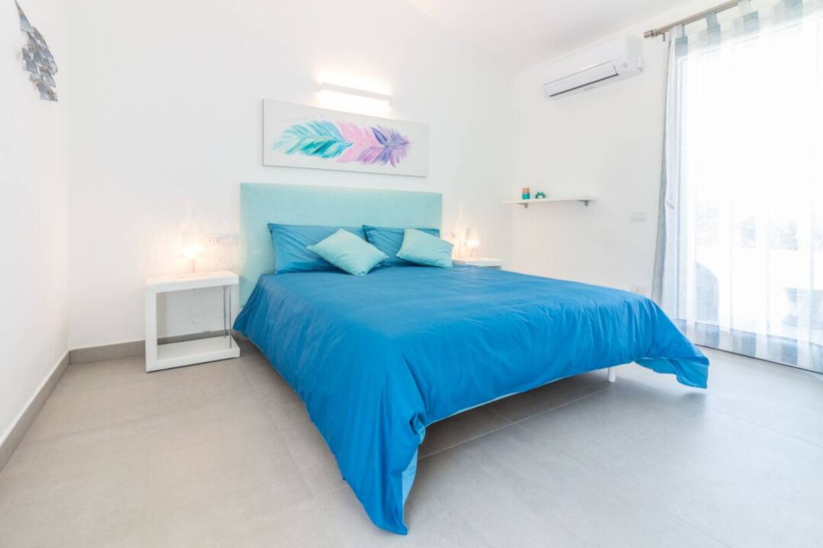 Een moderne slaapkamer met een groot bed, witte nachtkastjes, een blauw hoofdeinde en een groot raam met dunne gordijnen.