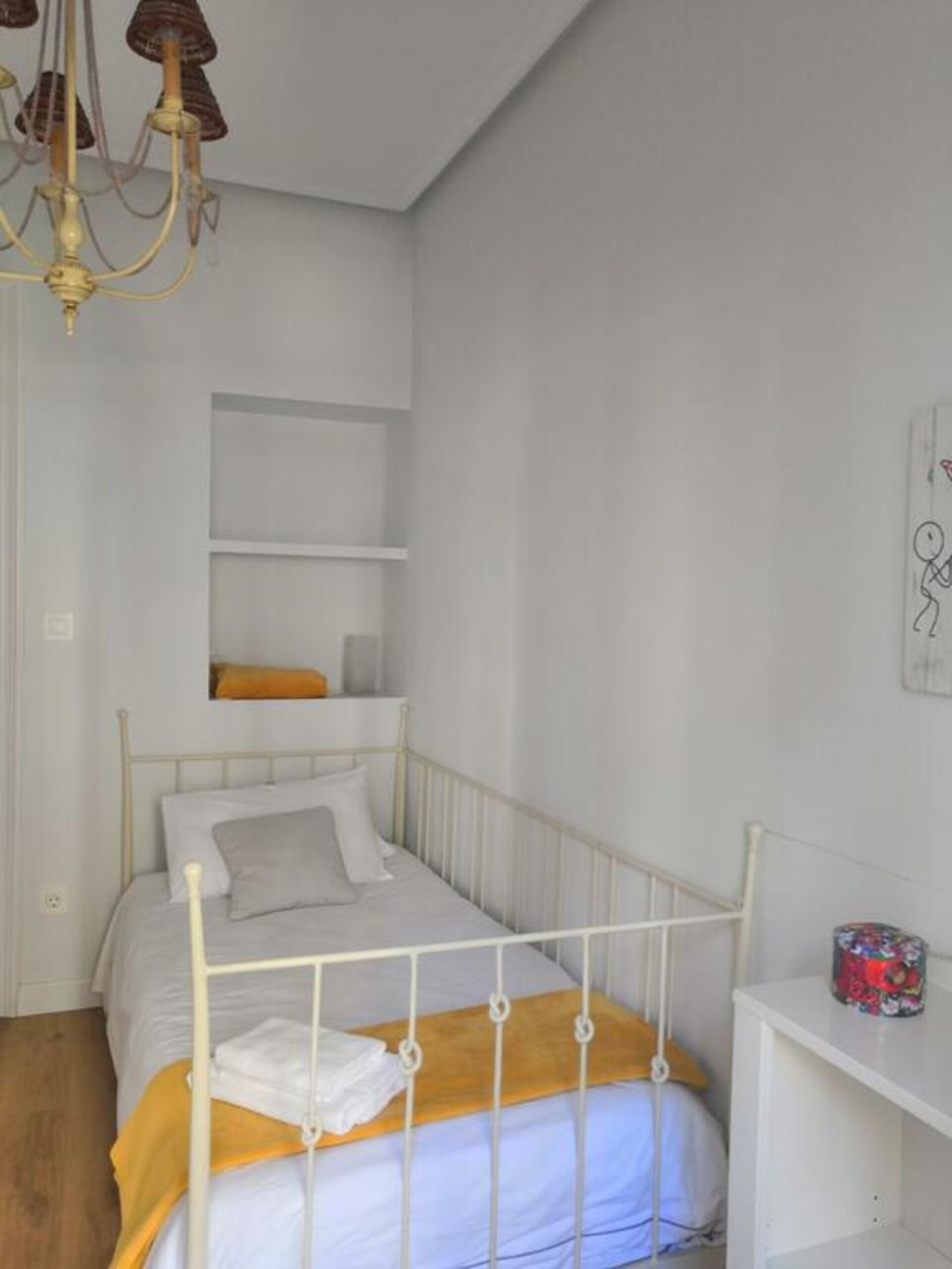 A single-bed room with a metal frame, white bedding, a wooden headboard, a chandelier, and a small shelf with a decorative item.