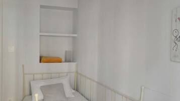A single-bed room with a metal frame, white bedding, a wooden headboard, a chandelier, and a small shelf with a decorative item.
