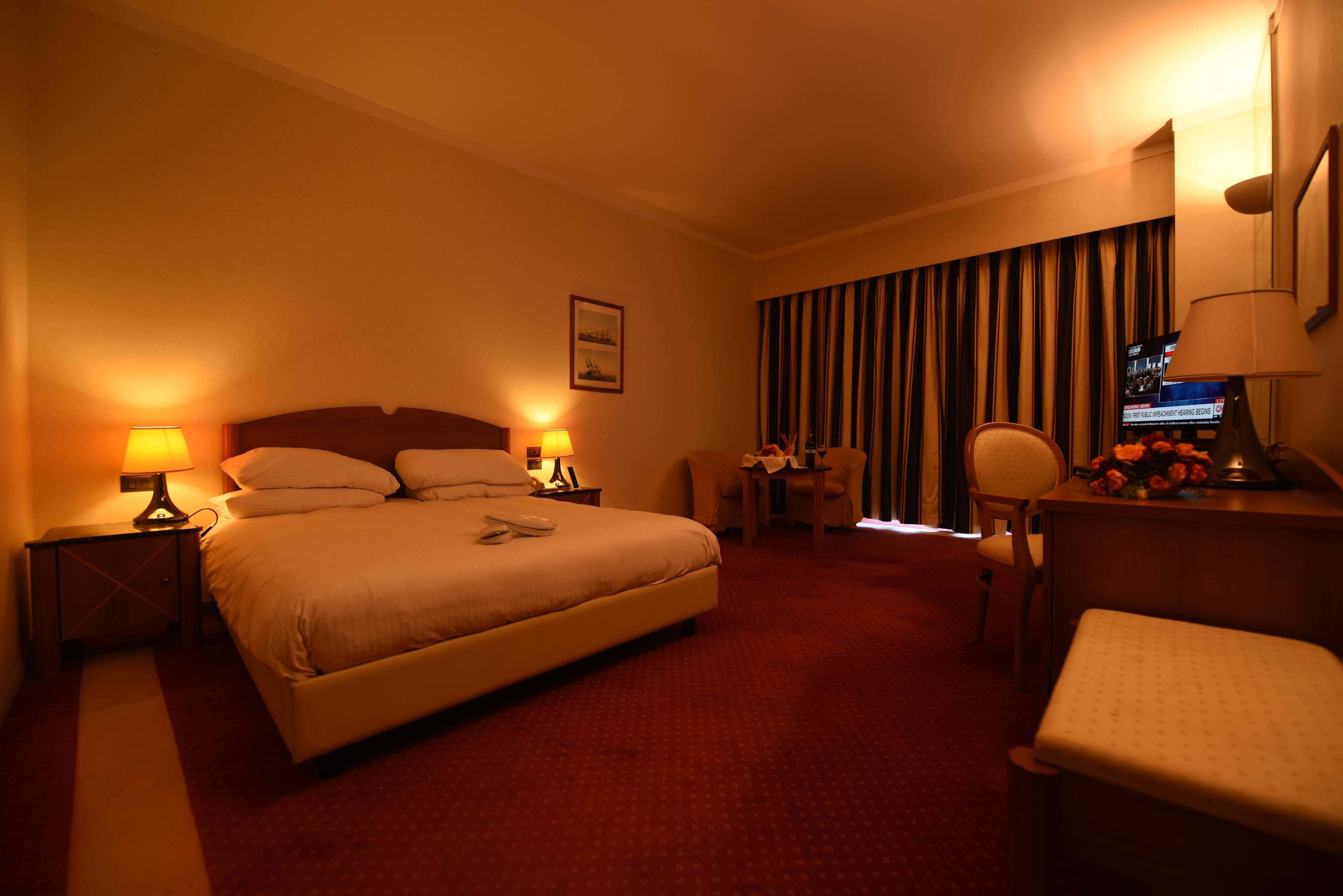 Ein Hotelzimmer mit einem großen Bett, zwei Nachttischlampen, einem Schreibtisch mit Stuhl, einem Fernseher und einem Fenster mit Vorhängen.