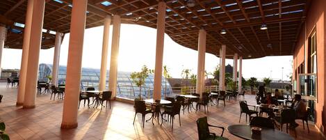 Uma área de café ao ar livre com mesas e cadeiras, pilares imponentes e vista para a paisagem urbana.