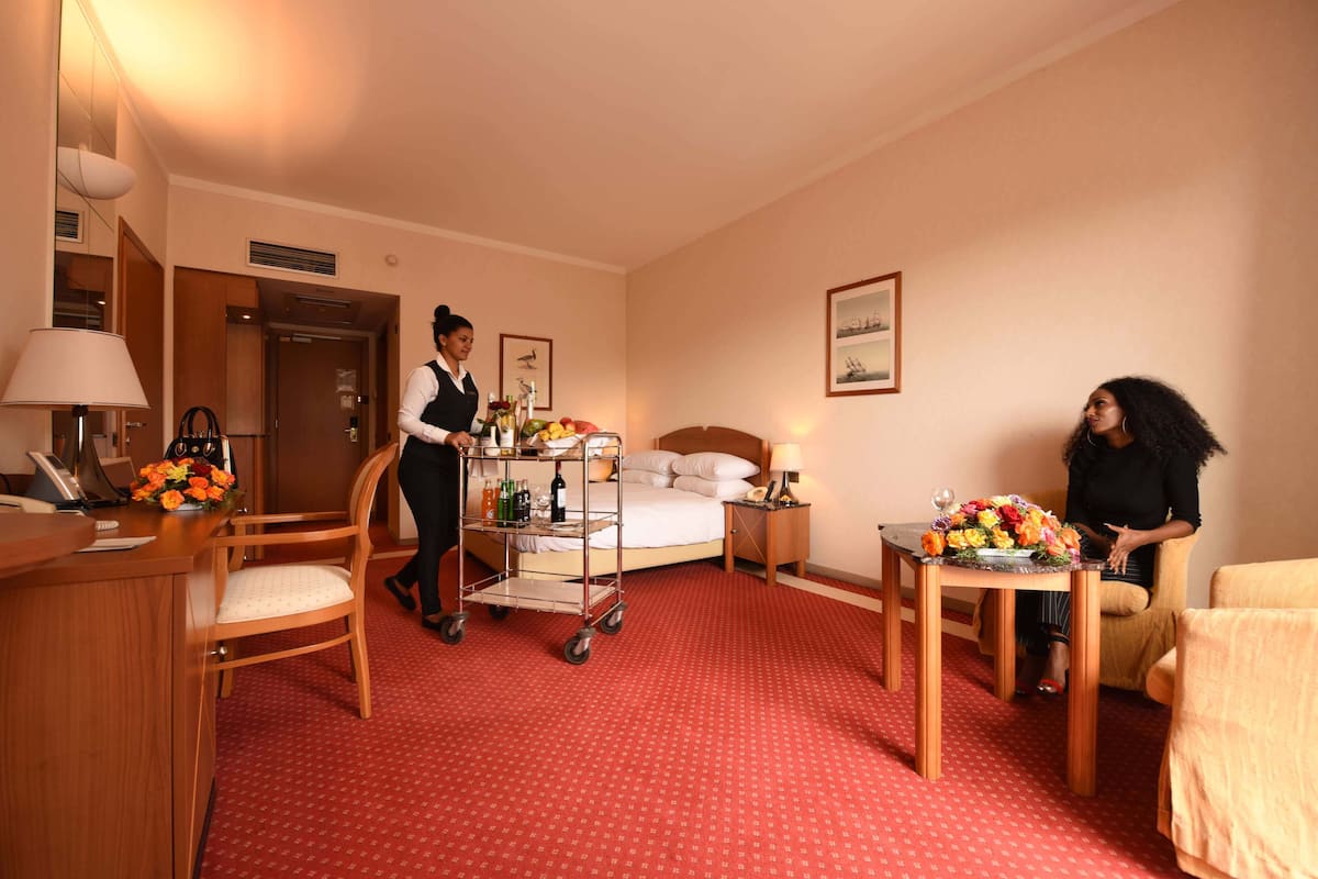 Ett hotellrum med en säng, ett skrivbord, en stol, ett litet bord med blommor, ett sängbord och en vägglampa.
