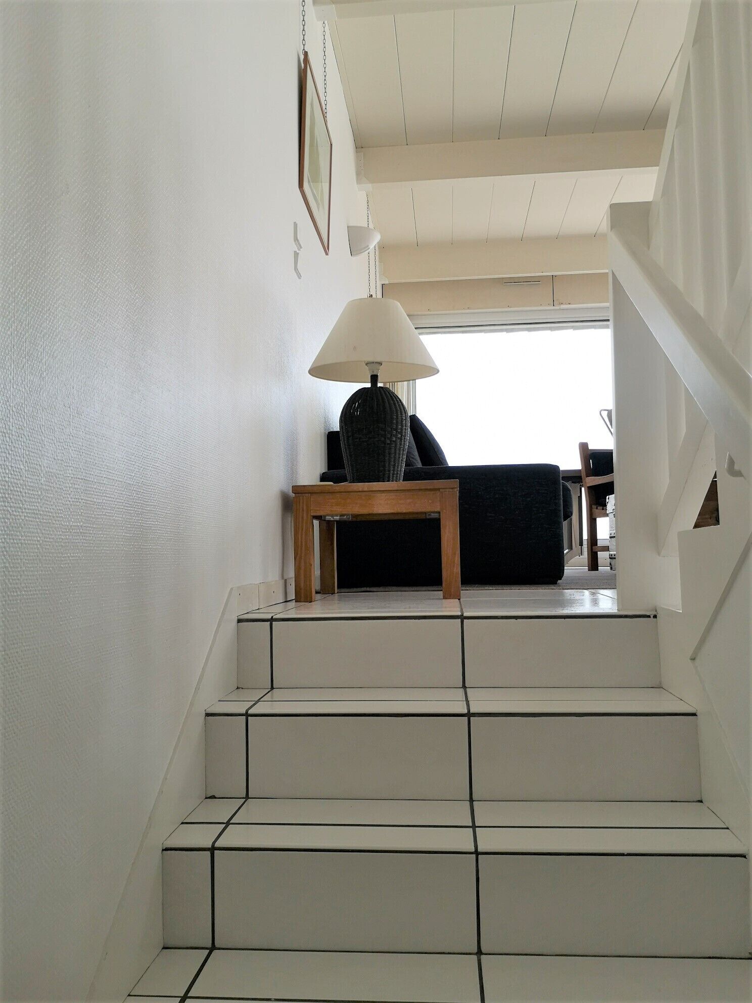 A staircase with white tiles leading to a living area with a lamp, sofa, and a framed picture on the wall.