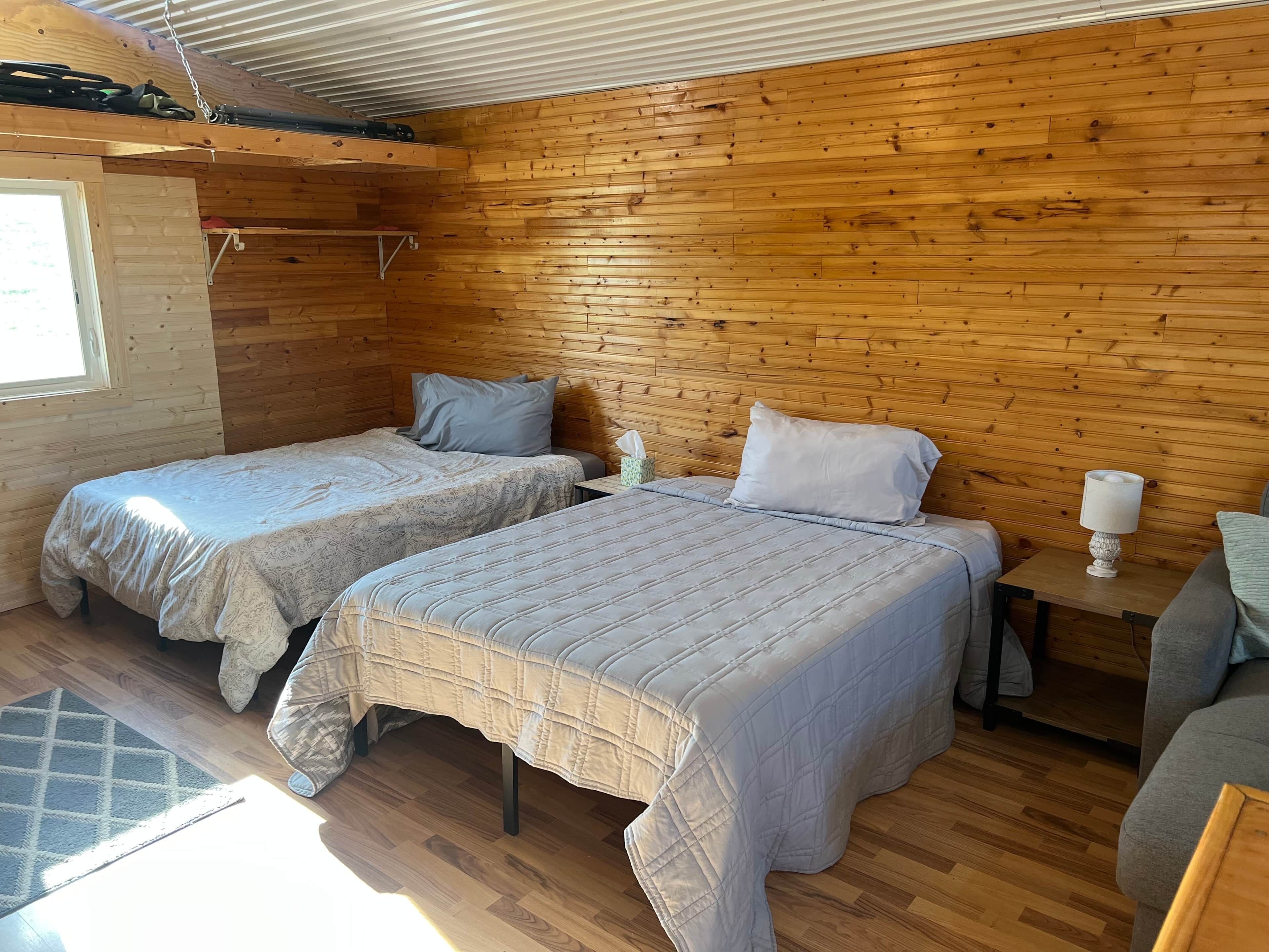 Acogedora habitación estilo cabaña con dos camas, paredes de madera, una mesita con una lámpara y una ventana.