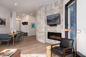 Un salon moderne avec une cheminée en pierre, un téléviseur fixé au mur et un coin repas avec des chaises bleues.