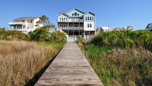 A two-story house with a wooden walkway leading to the entrance, surrounded by tall grasses and other vegetation. - Enjoy Sounds Beachy, an 8-bedroom haven in North Topsail Beach, NC with stunning water views. (North Topsail Beach)