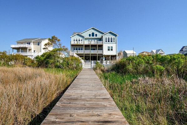 A two-story house with a wooden walkway leading to the entrance, surrounded by tall grasses and other vegetation. - Enjoy Sounds Beachy, an 8-bedroom haven in North Topsail Beach, NC with stunning water views. (North Topsail Beach)