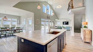 A spacious kitchen with a large island, a dining area with chairs, and a view of the beach through large windows. - Enjoy Sounds Beachy, an 8-bedroom haven in North Topsail Beach, NC with stunning water views. (North Topsail Beach)