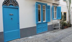 A building facade with a blue door and shutters, a metal bench, and potted plants.