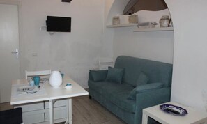 A small, modern room with a white table, a grey sofa, and a TV mounted on the wall. There is a shelf above the sofa with decorative items.
