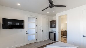 A bedroom with a bed, a ceiling fan, a TV mounted on the wall, and a kitchen area visible through an open door. - Poolside on the 2nd Green - Game Room, Sleeps 12 (St. George)