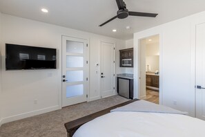 A bedroom with a bed, a ceiling fan, a TV mounted on the wall, and a kitchen area visible through an open door.