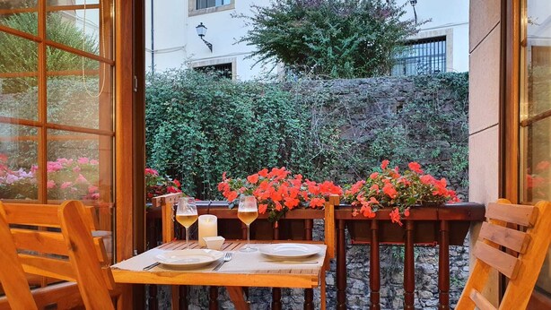 An outdoor dining area with wooden furniture and a table set for two, overlooking a garden with red flowers and a stone wall.
