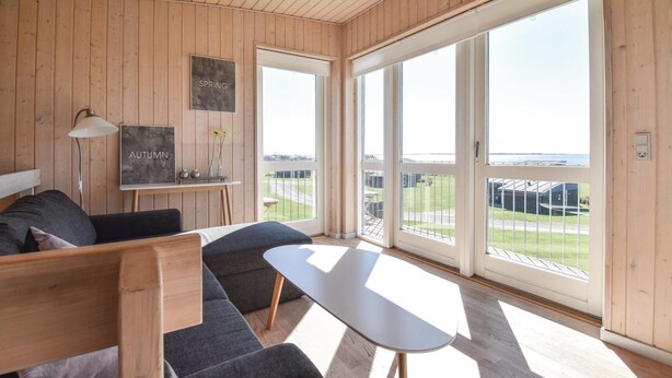 Une pièce moderne avec un intérieur en bois, un canapé, une petite table et de grandes fenêtres donnant sur l’extérieur.
