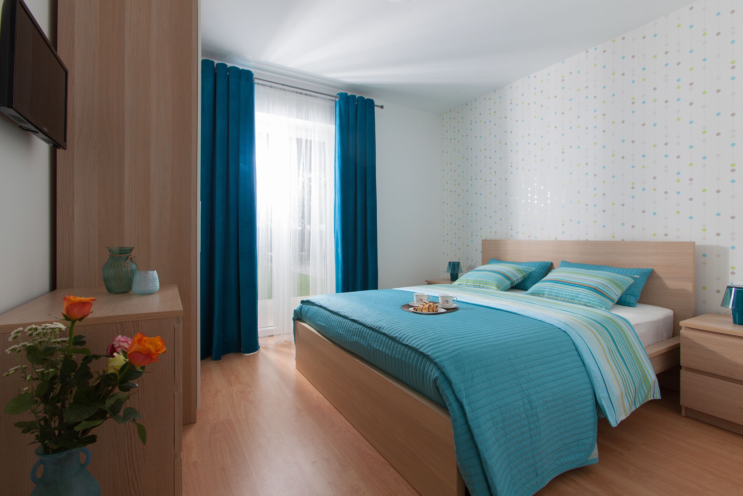 A bedroom with a bed, bedside tables, a nightstand, a vase with flowers, and a window with curtains.