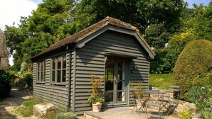 A small, single-story wooden cottage with a sloped roof, a front door, and a small porch area with a bench.