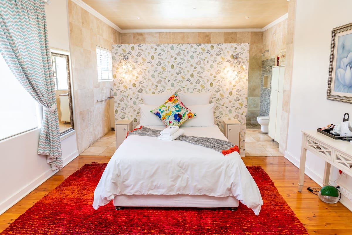 A bedroom with a large bed, a red and white rug, a floral-patterned headboard, and a white nightstand with a lamp.