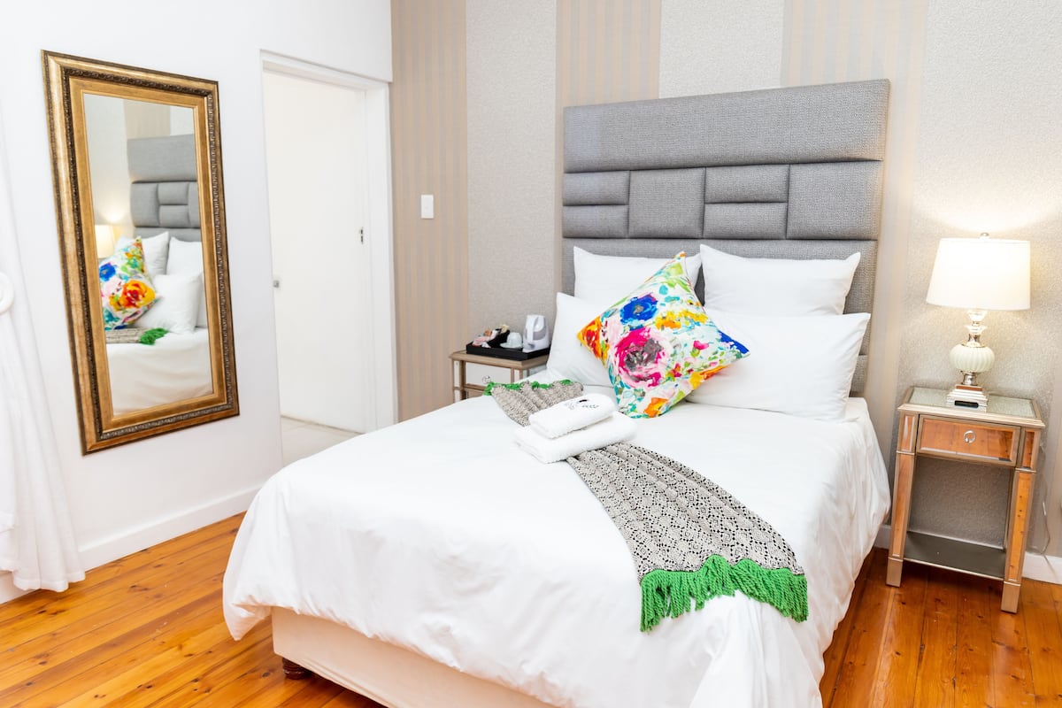 A neatly made bed with white linens, a grey upholstered headboard, and a floral pillow. There is a bedside table with a lamp and a small drawer. A mirror with a gold frame is on the wall to the left of the bed.