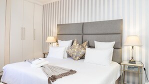 Een netjes opgemaakt bed met een grijs hoofdbord, wit beddengoed en decoratieve kussens. Er zijn twee nachtkastjes met lampen.