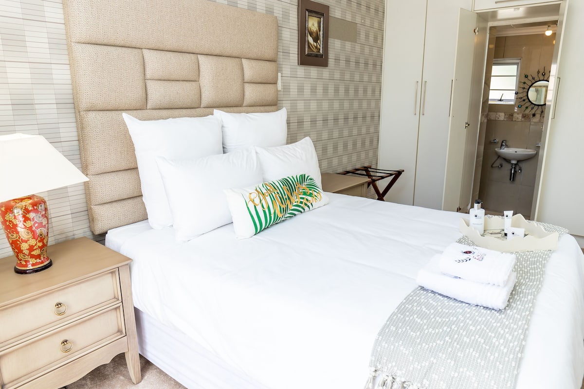 A neatly made bed with white linens, a headboard, and decorative pillows. A bedside table with a lamp and a decorative vase is visible.