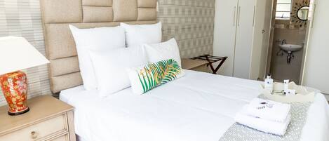 Una cama bien hecha con sábanas blancas, cabecera y almohadas decorativas. Se ve una mesita de noche con una lámpara y un jarrón ornamental.