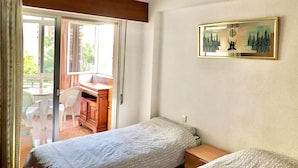 Quarto com duas camas de solteiro, uma escrivaninha de madeira, uma cadeira e uma janela com cortinas.