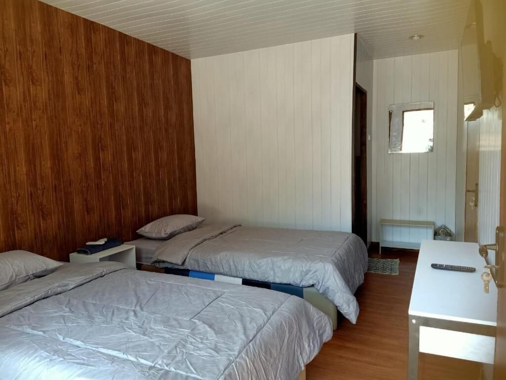 Une chambre avec deux lits, une tête de lit en bois, un mur blanc, une petite fenêtre et une table de chevet avec une télécommande.