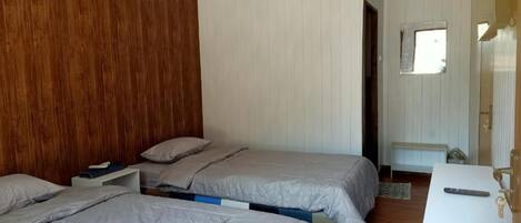 Une chambre avec deux lits, une tête de lit en bois, un mur blanc, une petite fenêtre et une table de chevet avec une télécommande.