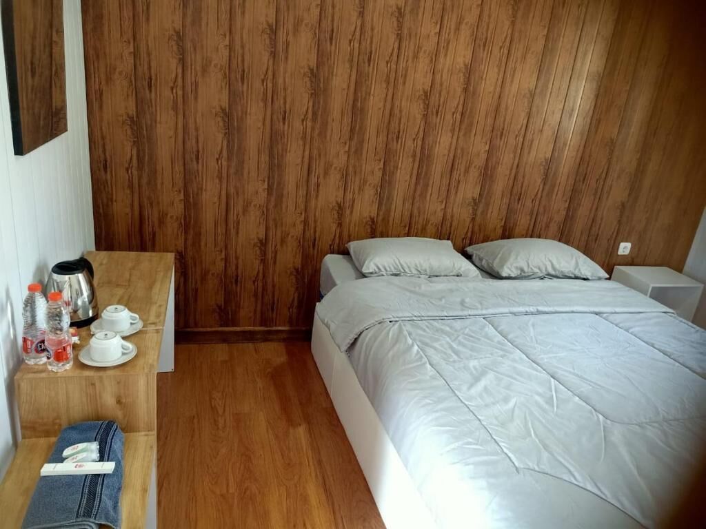 Una habitación con pared de madera, una cama con ropa de cama blanca, una mesita de noche con hervidor, tazas y una botella.